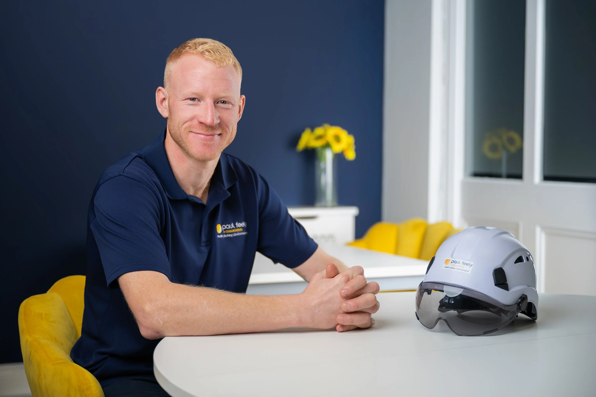 A man with ginger hair and a beard, wearing a blue polo shirt with the logo 'Paul Feely' sits at a white table with a white construction helmet in front of him, in a brightly lit room with yellow chairs, a dark blue wall, and yellow flowers in a vase.