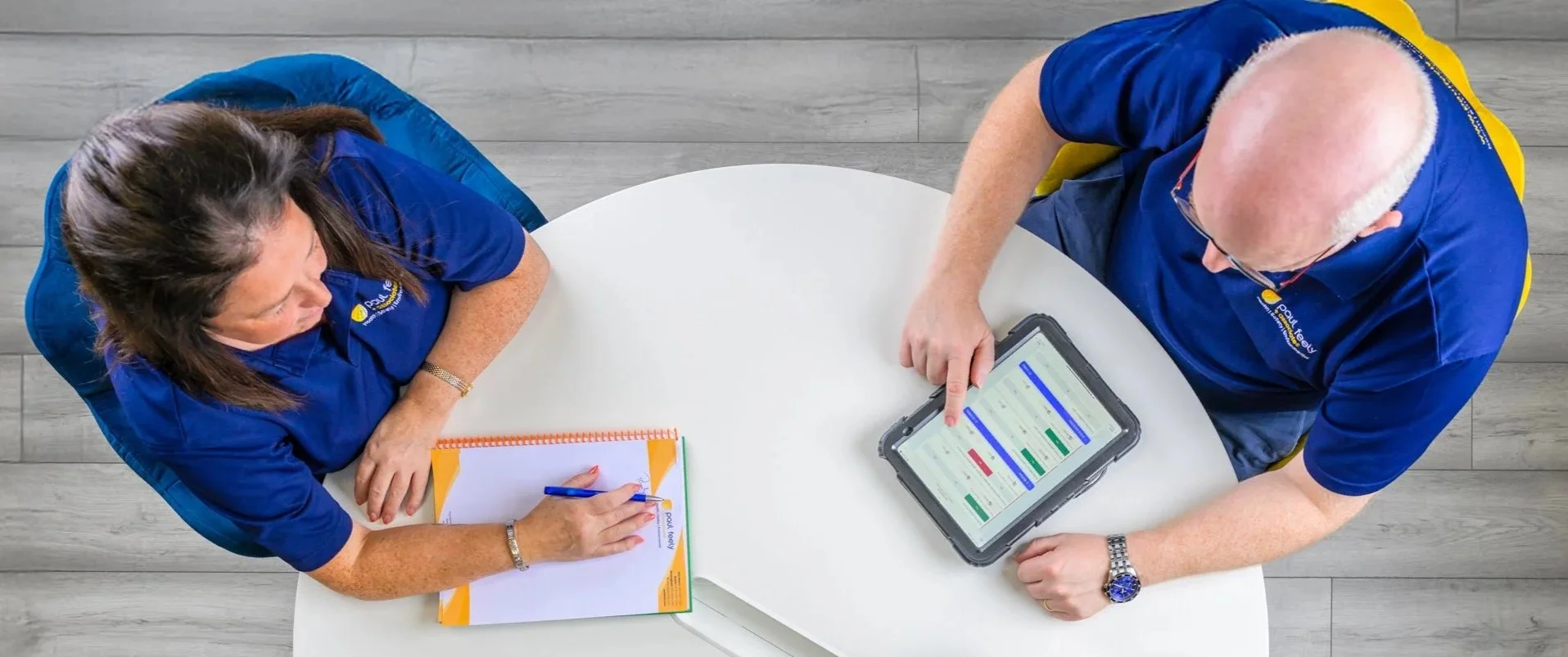 Two people in blue shirts sitting at a round white table, discussing work. One person is using a tablet with a scheduling app, and the other has a notepad and pen.