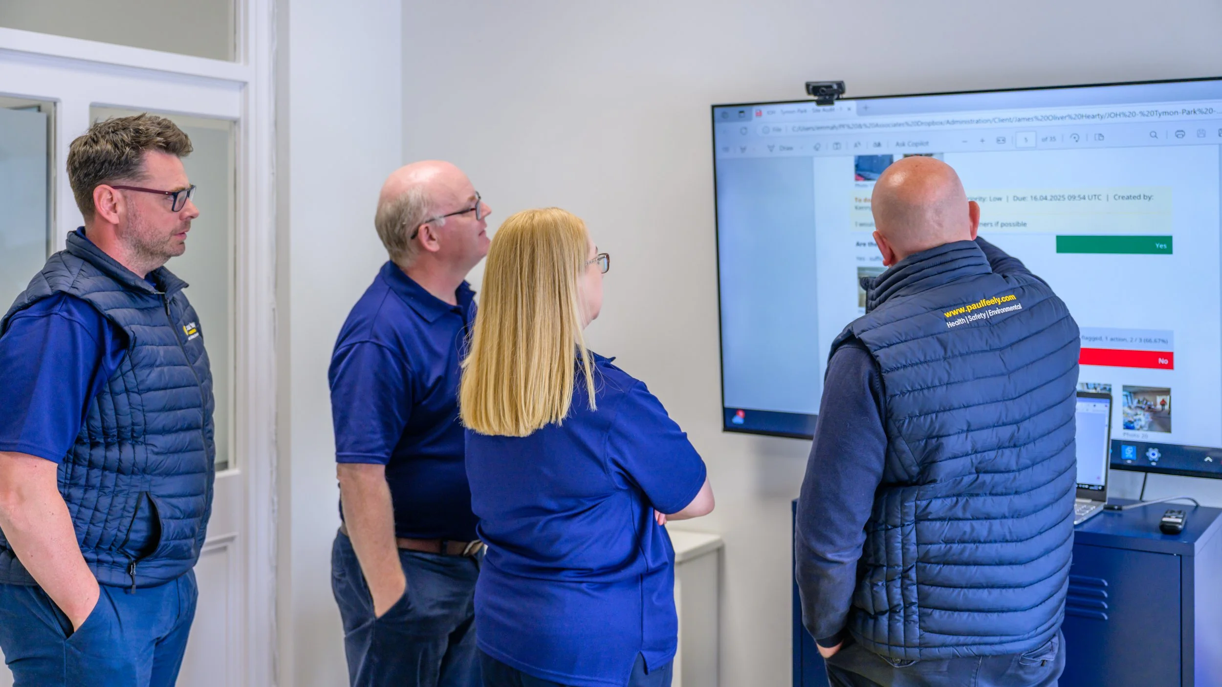 Four individuals in blue and black jackets and a woman in blue scrubs, standing and looking at a large screen showing a survey or form with checkmarks and text.