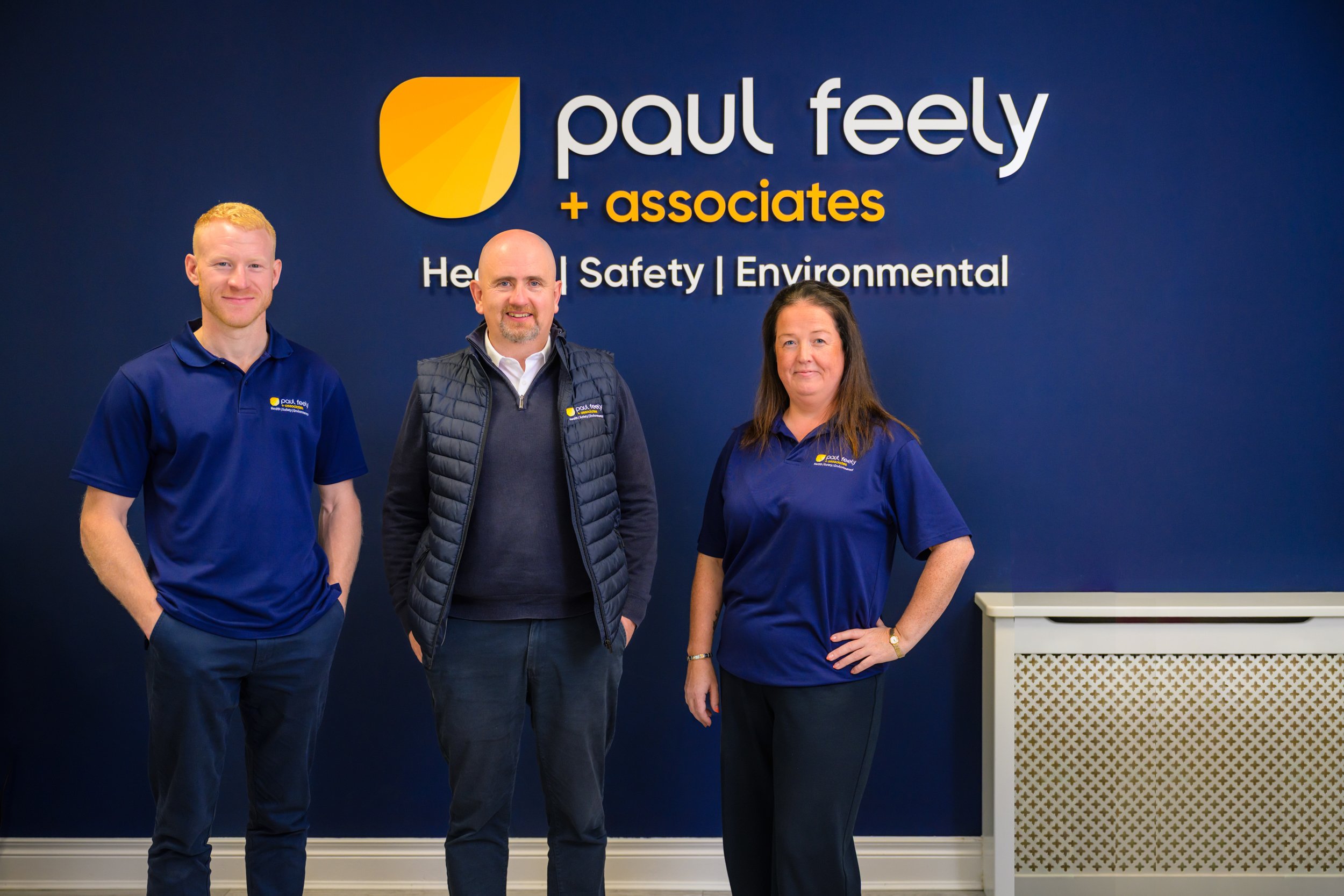 Three staff members standing in front of a large sign that says 'Paul Feely + associates' with the words 'Health | Safety | Environmental' underneath. They are wearing blue shirts or jackets with the company logo, and are in an office or corporate setting.
