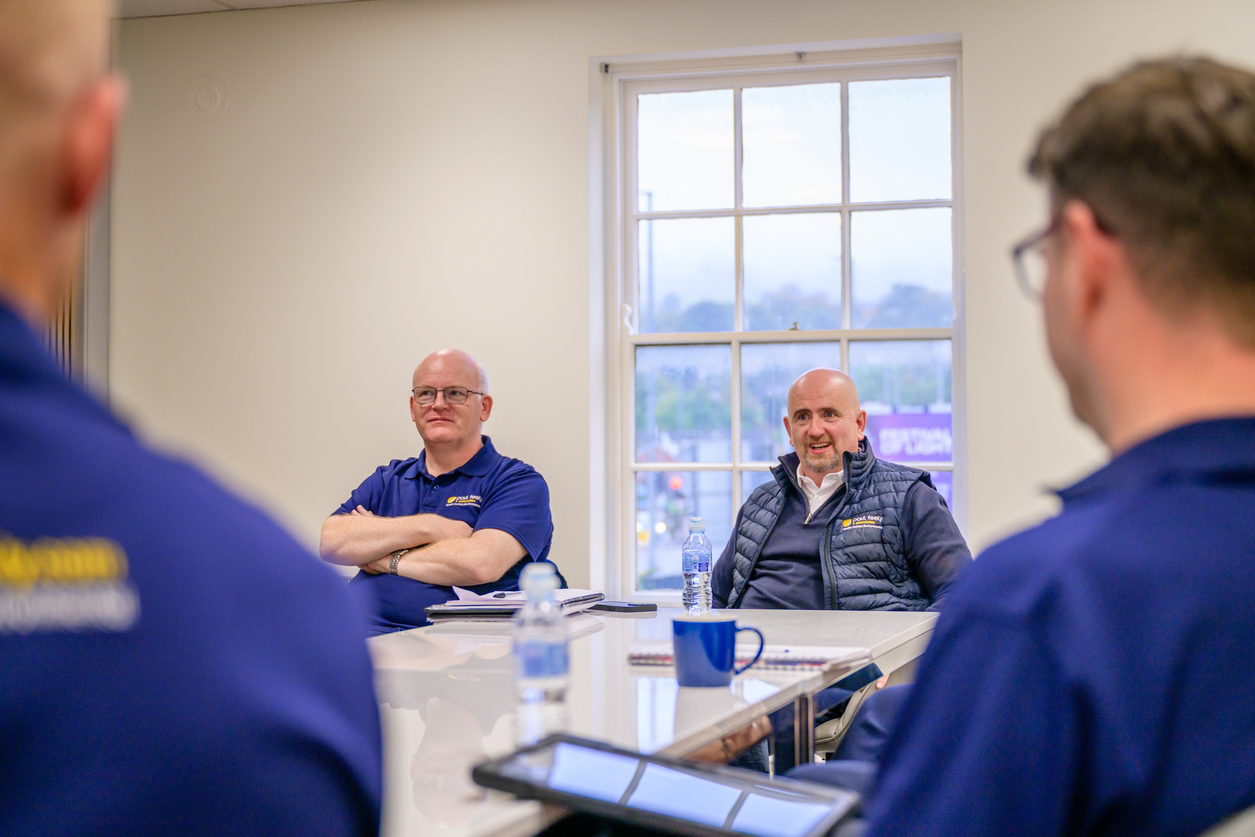 A group of men in a meeting room, some wearing company uniforms, sitting around a table. Two men are clearly visible, one bald man with glasses and another bald man wearing a vest, both engaging in conversation or listening, with water bottles, noteb