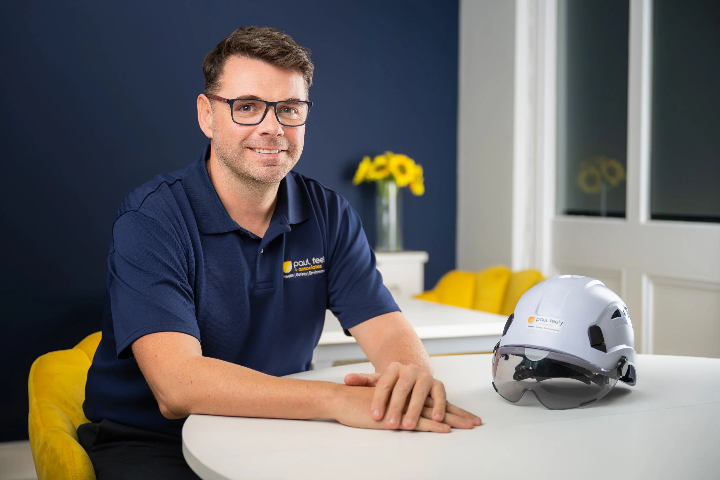A smiling man wearing glasses and a navy polo shirt sitting at a white table in a bright room with a dark blue wall, yellow chairs, a vase with yellow flowers, and a white helmet on the table.