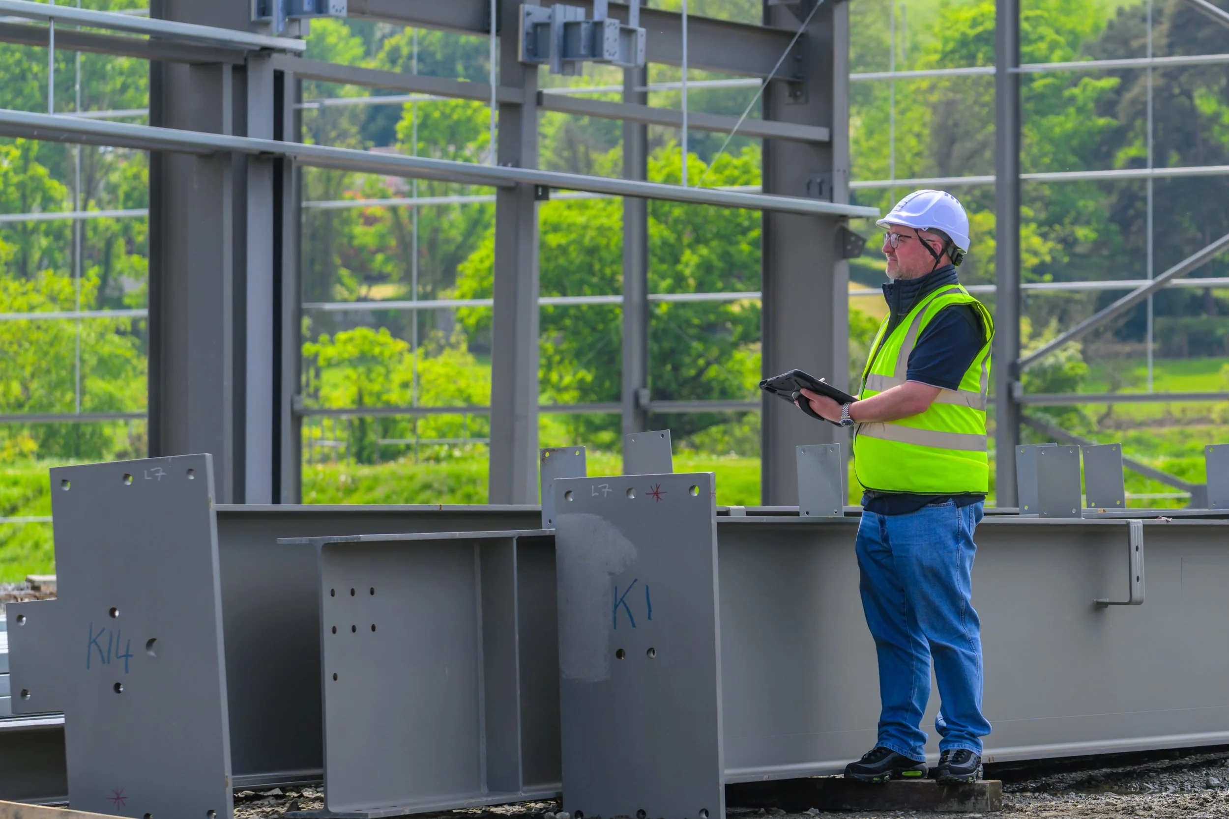 A construction worker wearing a yellow safety vest, white helmet, and glasses holding a tablet, standing on a construction site with metal framework and green trees in the background.