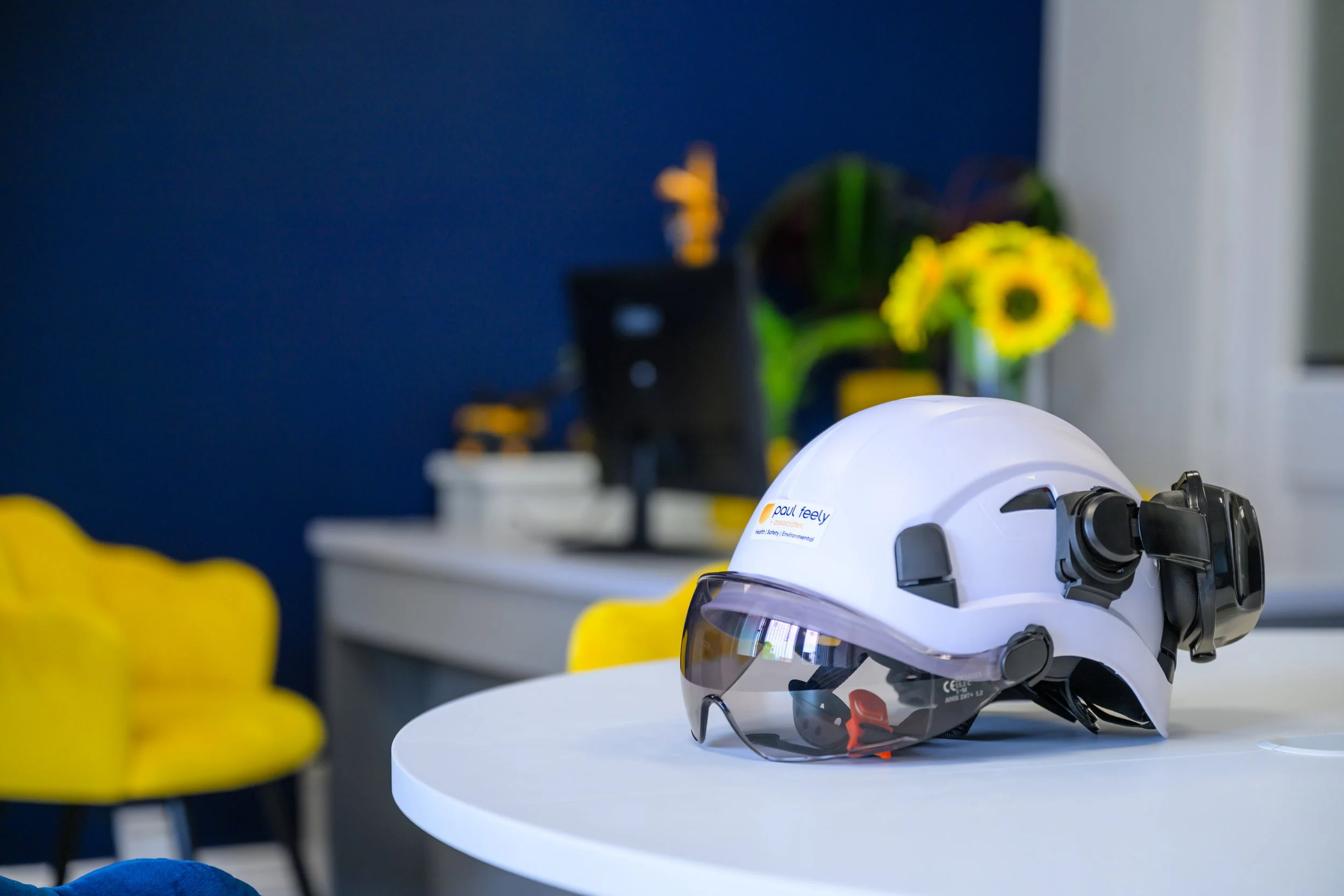 A white safety helmet with attached goggles and a camera, placed on a white table in a modern office or workspace with yellow chairs, a dark blue wall, a white desk, a computer monitor, and a vase of yellow sunflowers in the background.