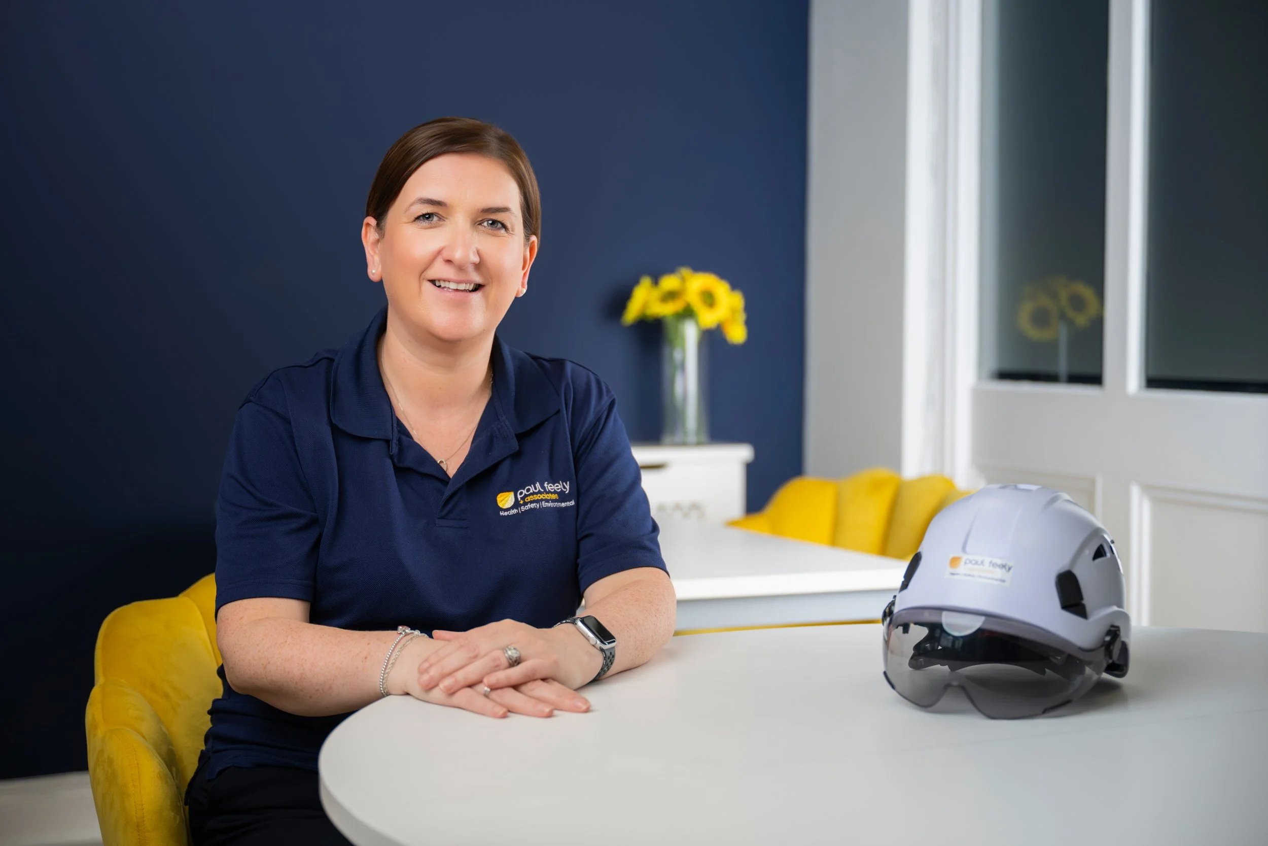 A woman sitting at a white table with a helmet on it, in a room with a navy blue wall, a yellow chair, and a vase of yellow flowers in the background.