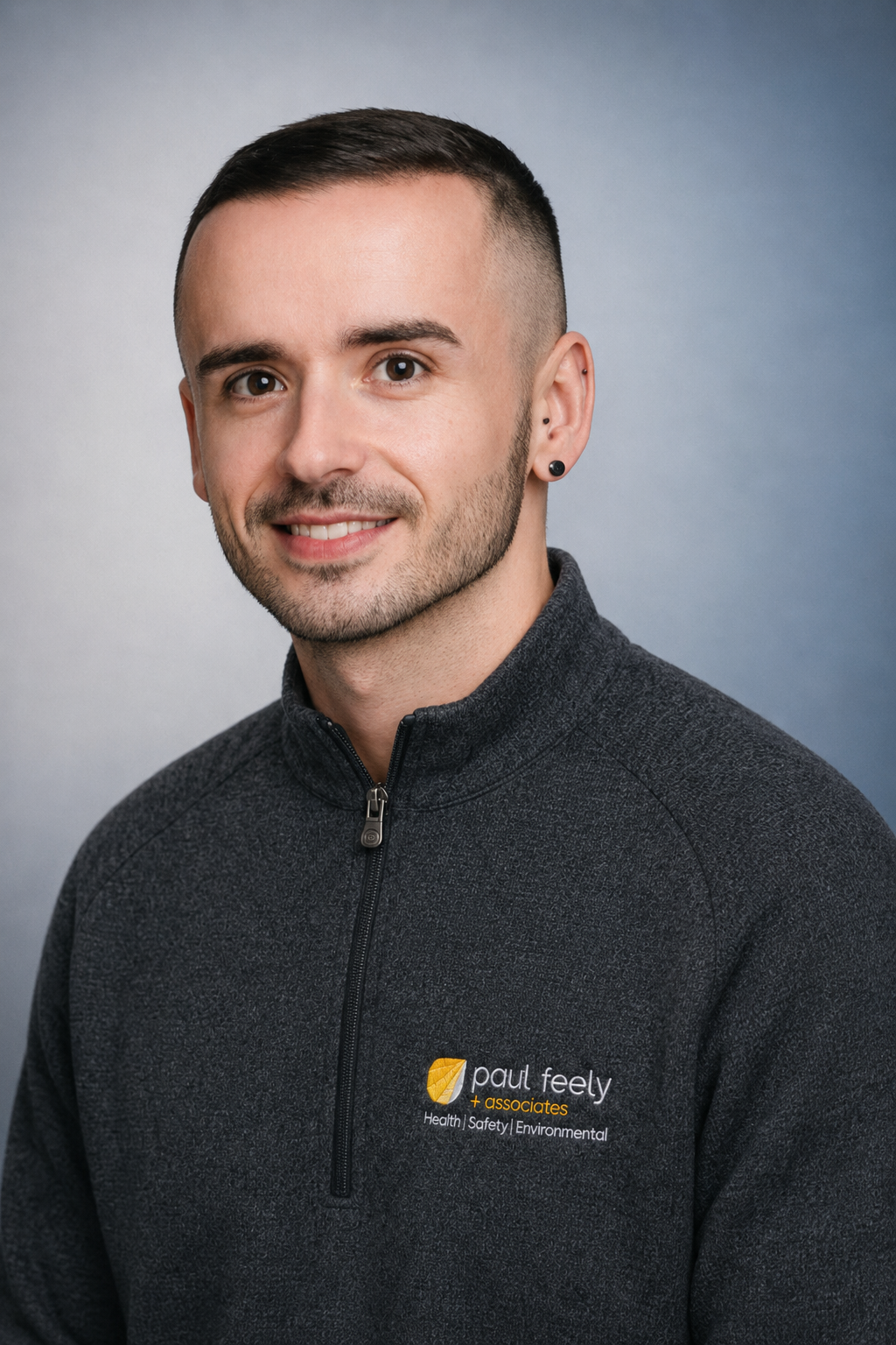 Portrait of a young man with short dark hair and a goatee, smiling, wearing a dark gray zip-up athletic jacket with a logo reading 'Paul Feely + associates, Health Safety Environmental' on it, against a plain light gray background.