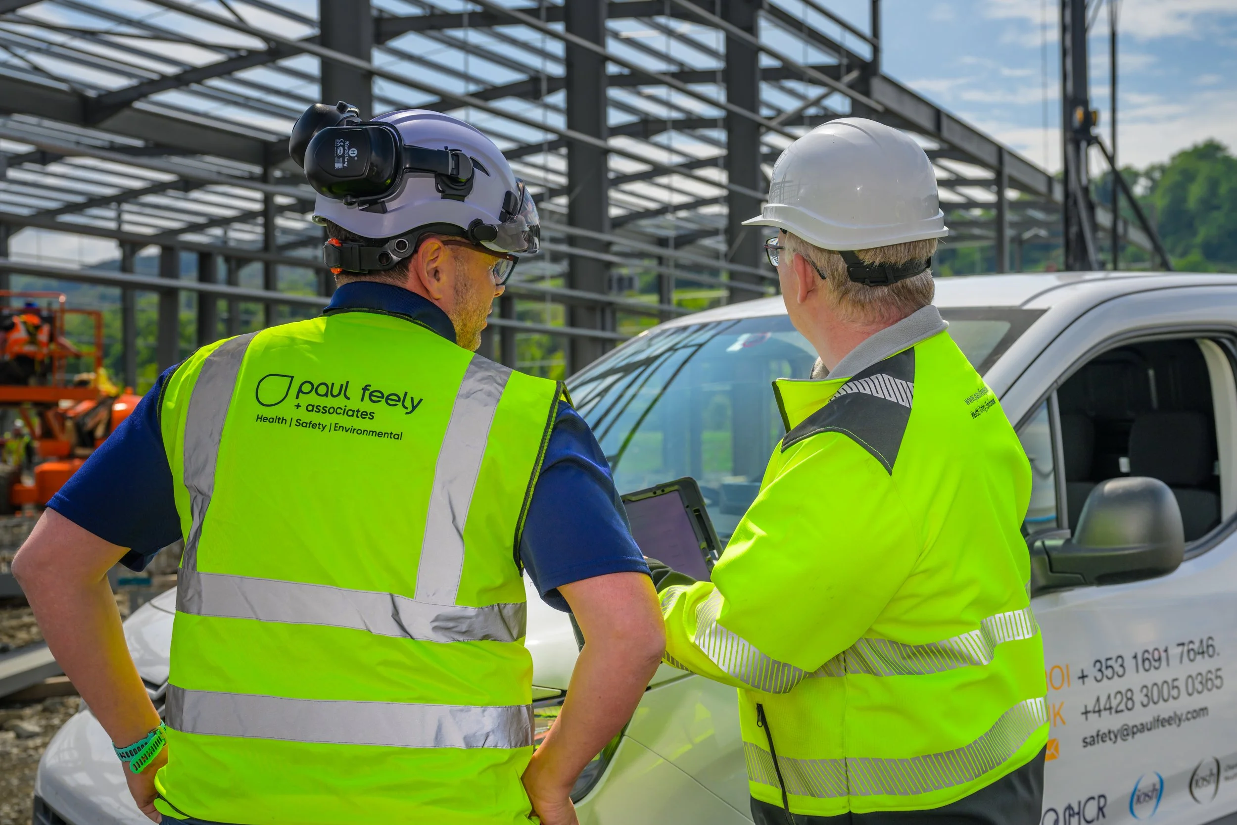 Two construction workers wearing yellow safety vests and hard hats inspecting documents near a white vehicle at a construction site.