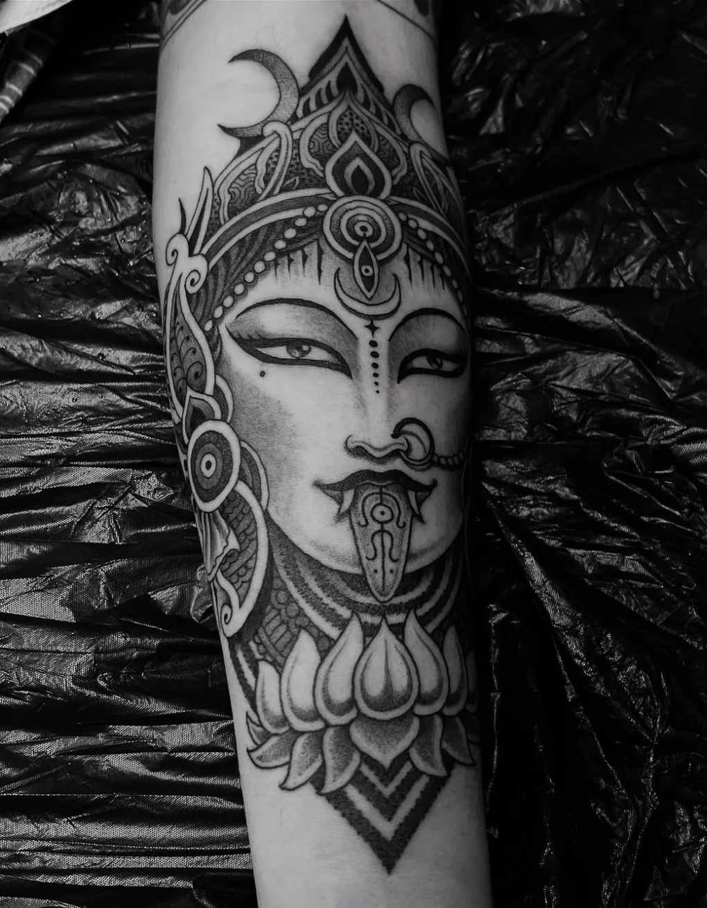 A black and gray tattoo of a serene feminine face with traditional ornamental details, a nose ring, and a lotus flower below, on a person's arm.