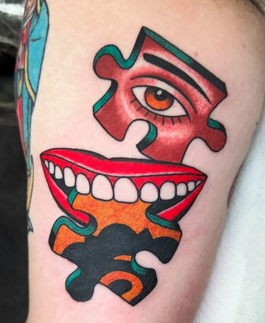 Tattoo of a puzzle piece with an eye and a mouth, featuring bold colors and outlines.