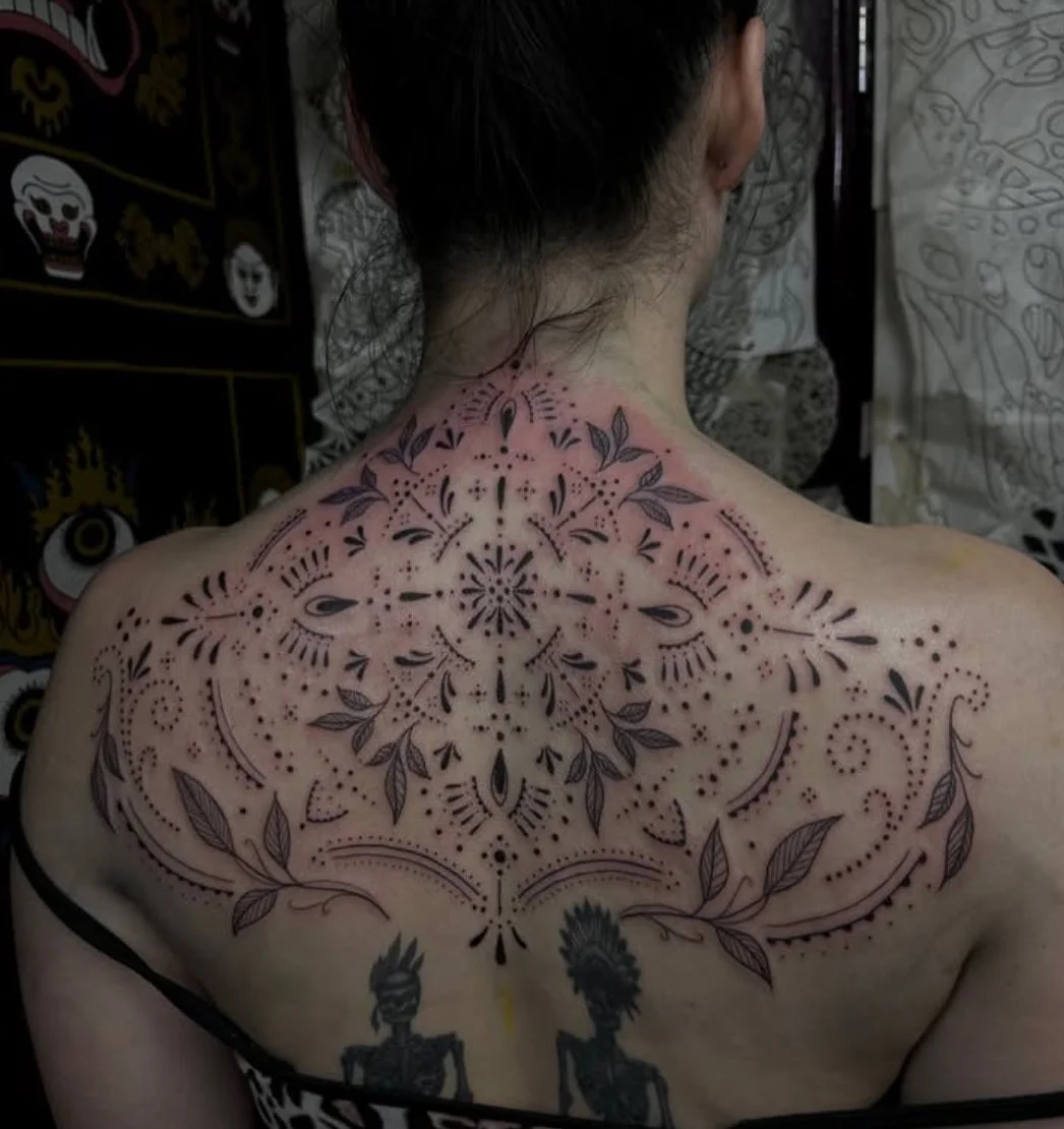 Back of a person with a large, intricate black tattoo design featuring floral and geometric patterns extending across the shoulders and upper back.