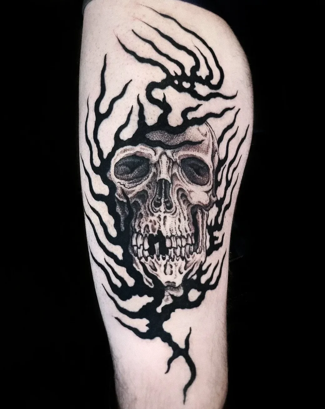 Black and gray tattoo of a skull with hollow eyes and detailed bones, surrounded by black abstract lines and shapes.