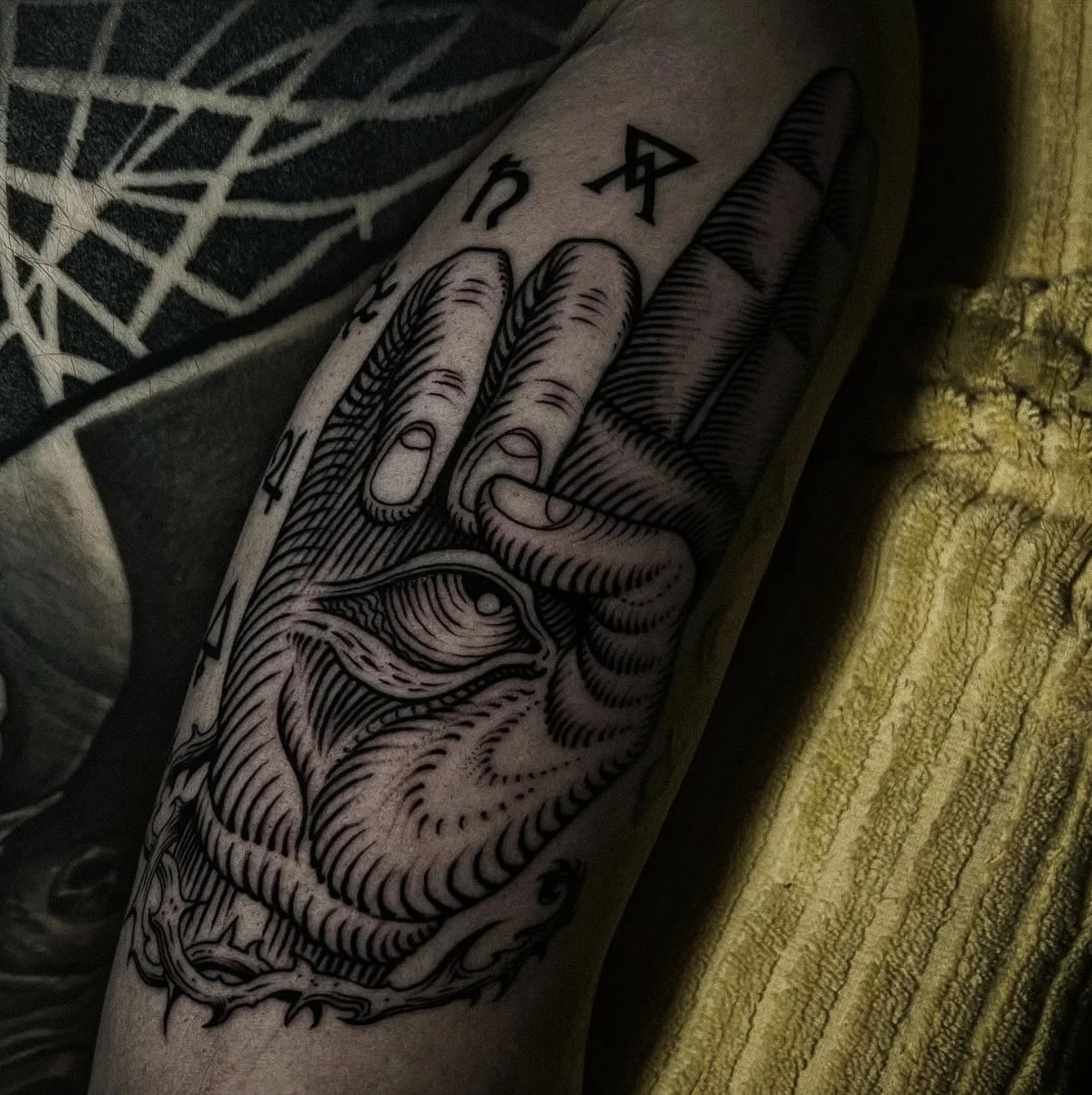 Black and gray tattoo of a hand with an eye in the palm, making a peace sign, on a person's arm.