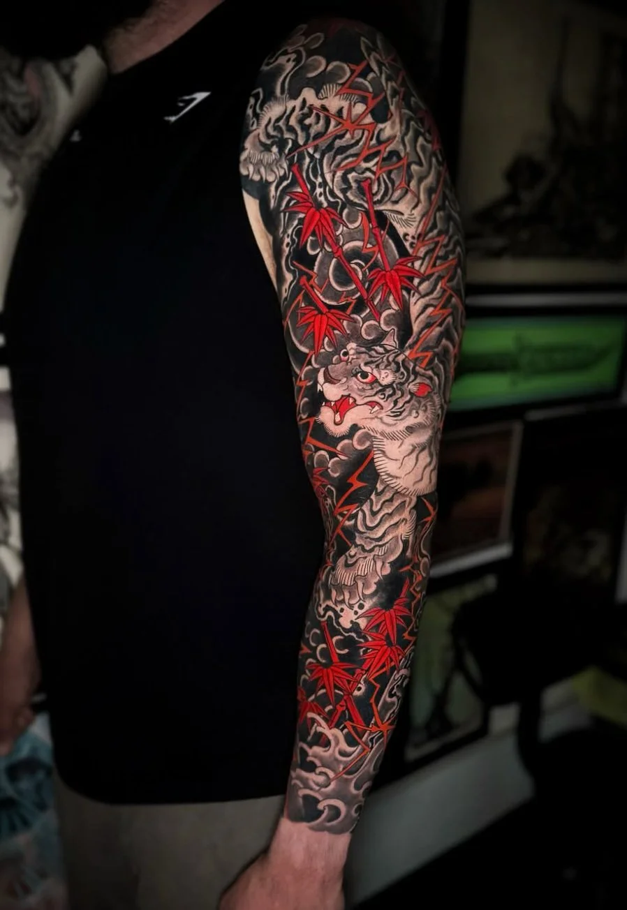 Tattoo of a fierce tiger face with red eyes and sharp teeth, surrounded by red maple leaves and black storm-like clouds on a person's upper arm.