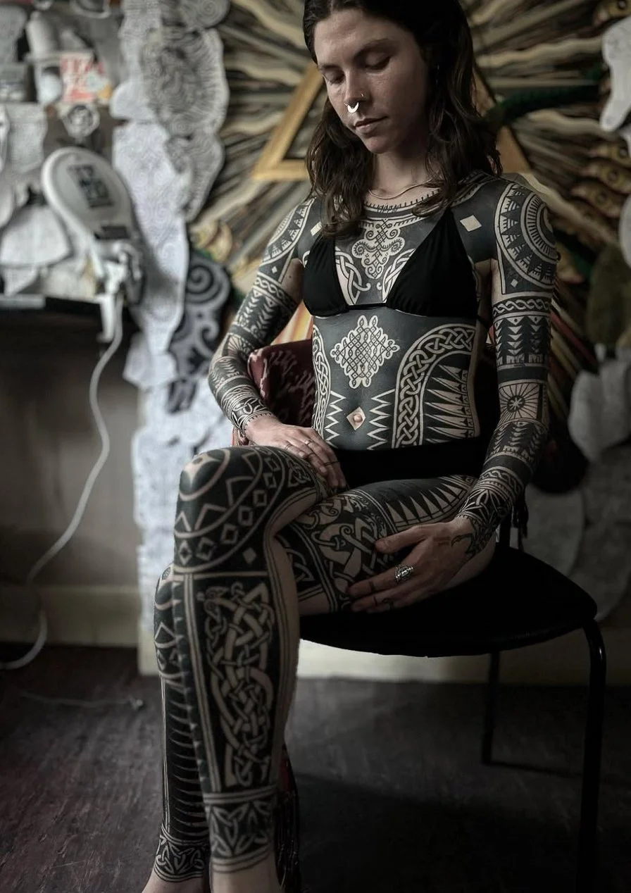 A young woman with long dark hair and tattoos covering her arms, torso, and legs, sitting on a black chair in a tattoo studio, wearing a black bikini top. The studio has artwork and tattoo designs on the wall behind her.