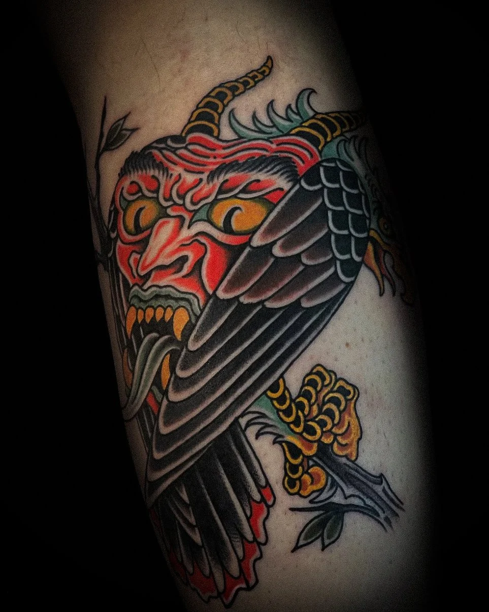 Colorful traditional tattoo of a fierce face with dragon-like features, a red and black wing, and other decorative elements.