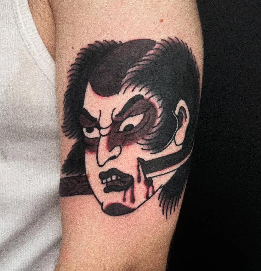 A tattoo of a stylized, aggressive man's face with dark hair, eyebrows, and facial features, in a bold, graphic style. The man is biting a knife, and red ink depicts blood dripping from his mouth and the knife.