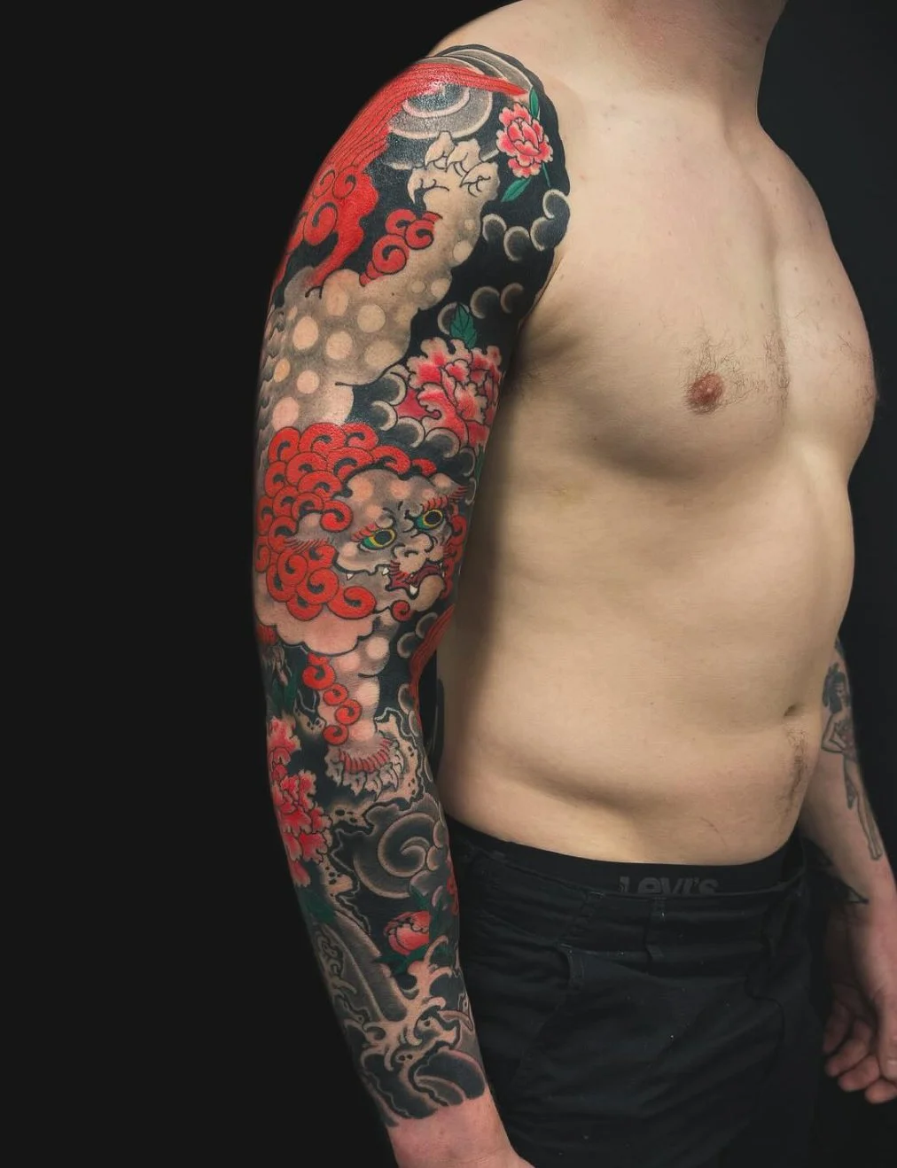 A shirtless person showing a colorful Japanese-style tattoo sleeve on their left arm, featuring a dragon, clouds, and floral elements.
