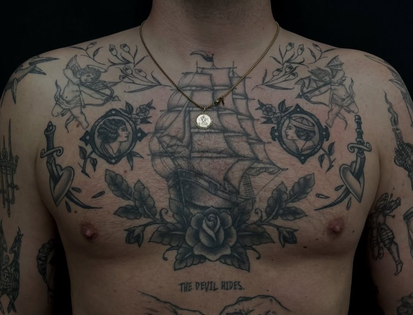 Tattooed chest with a ship, floral elements, portraits, and swords, with a necklace featuring a circular medallion.