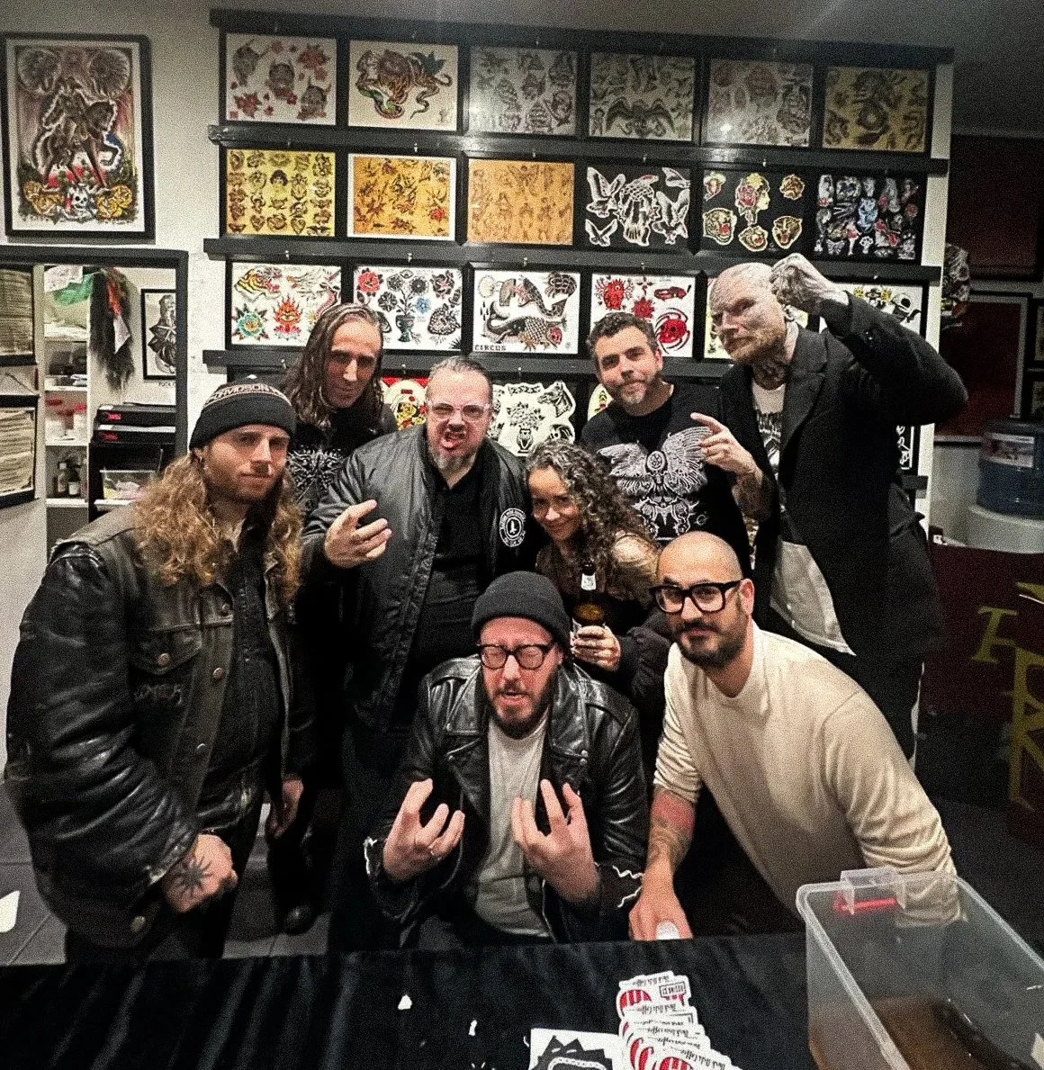 Group of eight people posing together in a tattoo shop behind a table with tattoo parlor artwork on the wall.