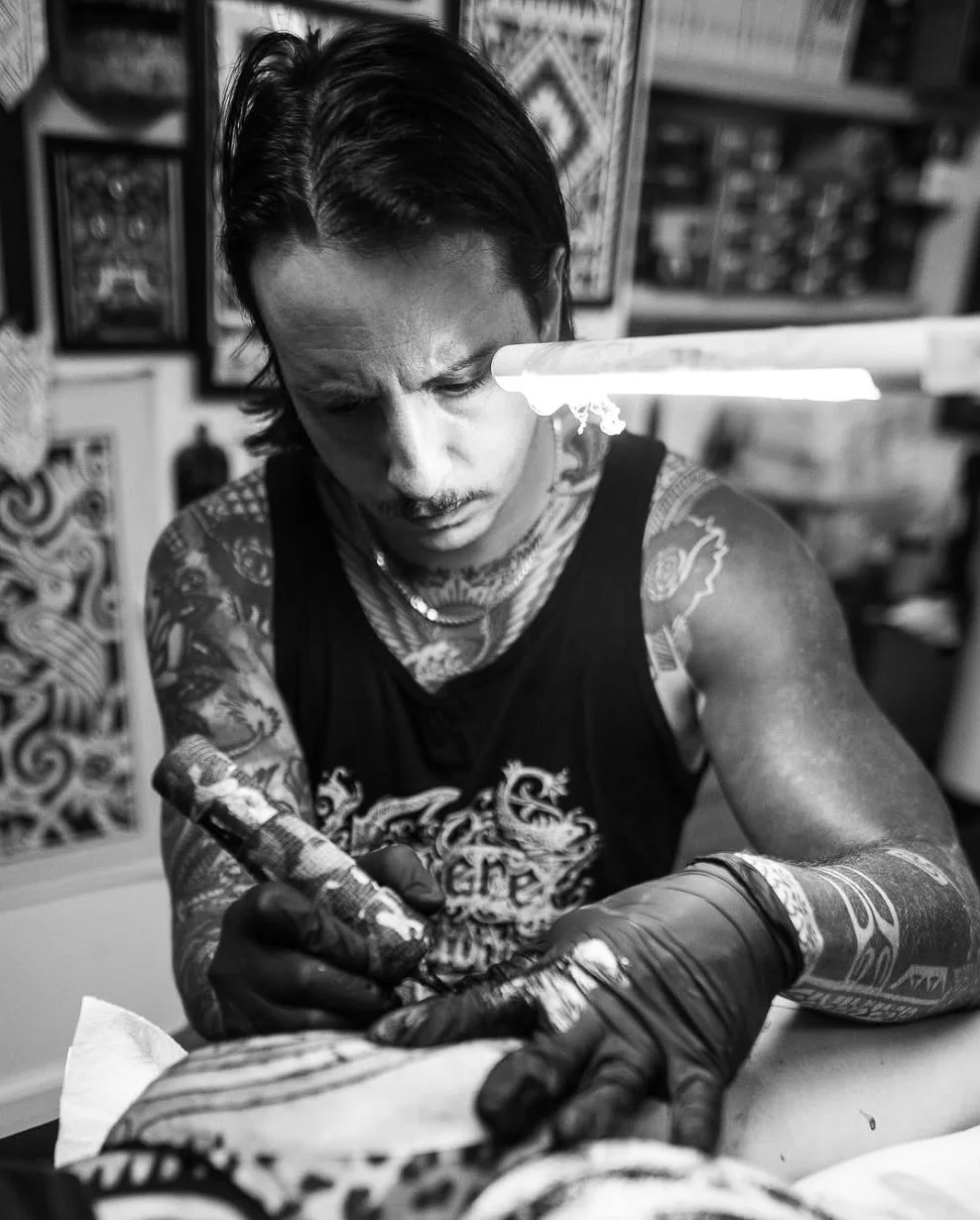 A tattooed man with long hair and a focused expression, wearing a sleeveless shirt, is tattooing skin with a tattoo machine in a tattoo studio.