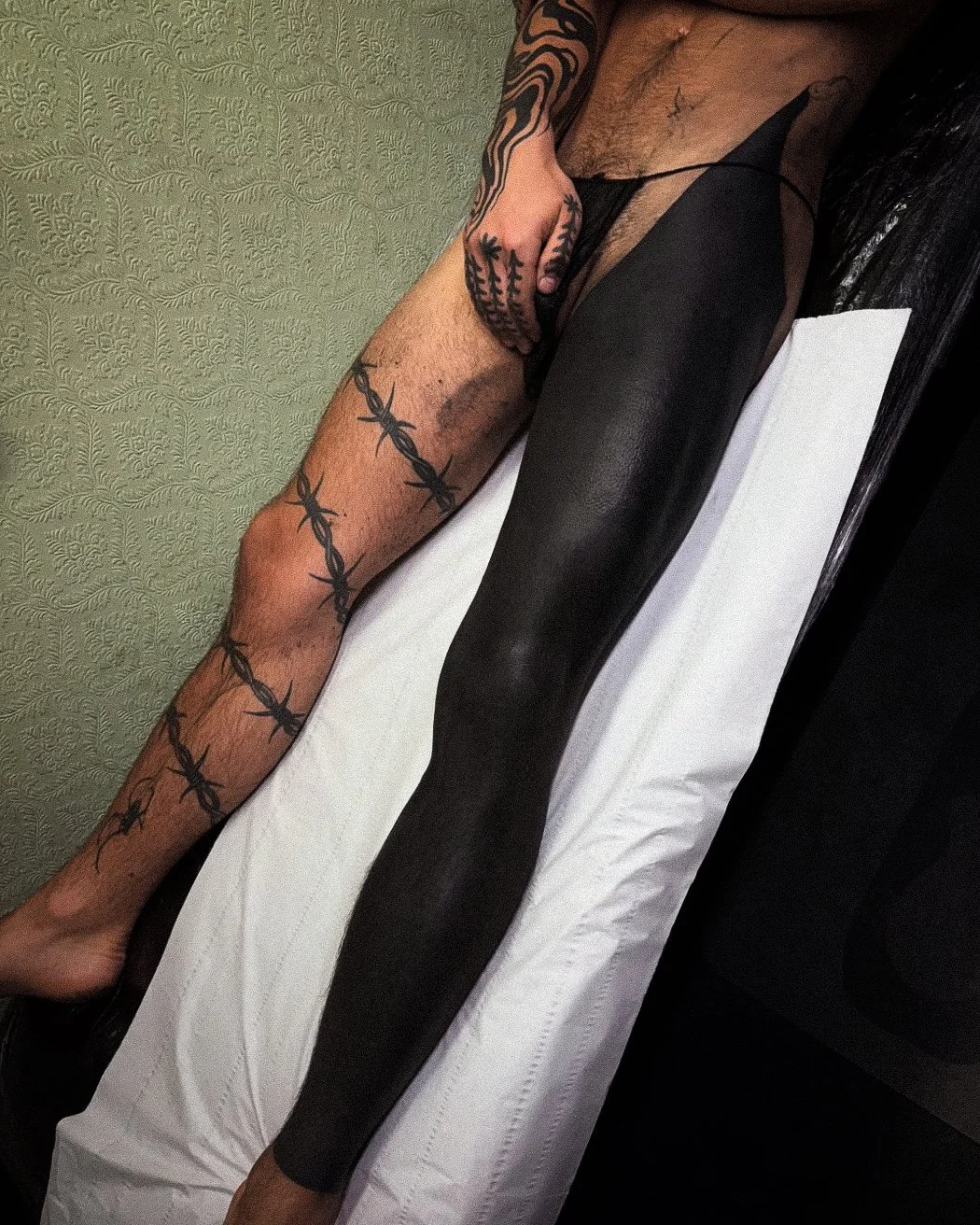 A person with numerous tattoos on their arm and hand, lying on a white and black surface, with their arm extended and hand resting near their head, wearing black sheer clothing under black and white sheets.