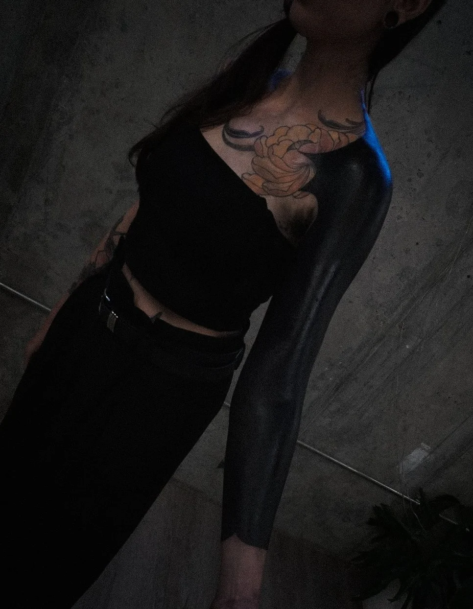 A woman with a large tattoo on her chest and shoulder, wearing a black crop top and black pants, standing in a dimly lit room with gray walls.