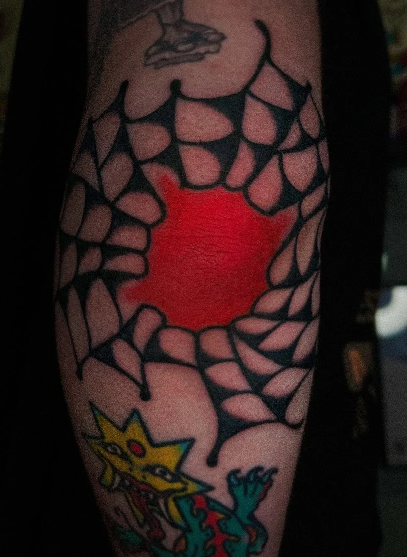 Tattoo of a black spider web with a red center and a cartoonish yellow cat with green tail and red details.