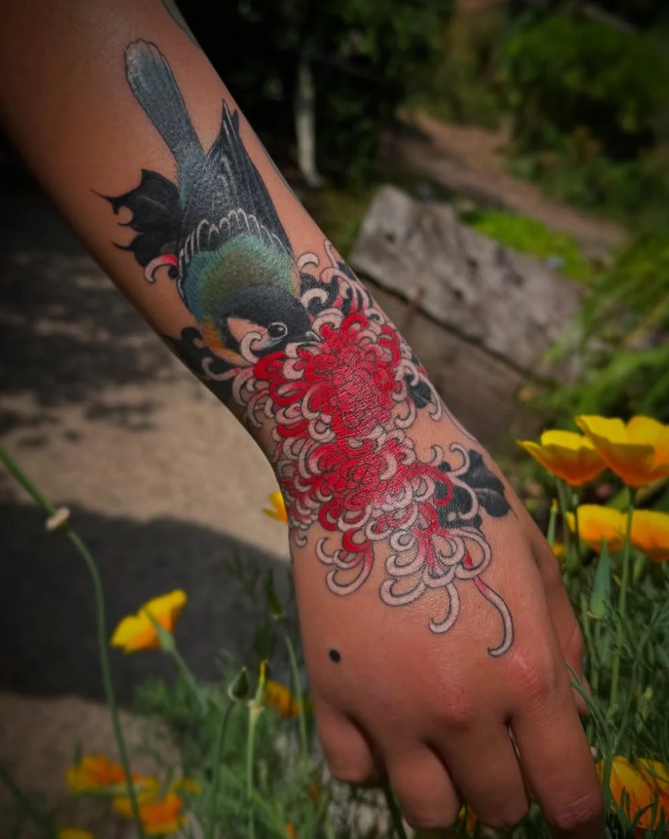 A person's arm with a colorful tattoo of a bird and red flowers, surrounded by yellow flowers in the background.