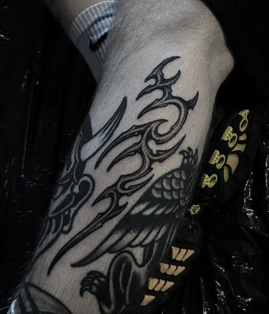 Close-up of a person's arm with a black tribal-style tattoo featuring a wing and intricate swirling patterns, wearing a gray Nike cap and black clothing with yellow text and numbers.