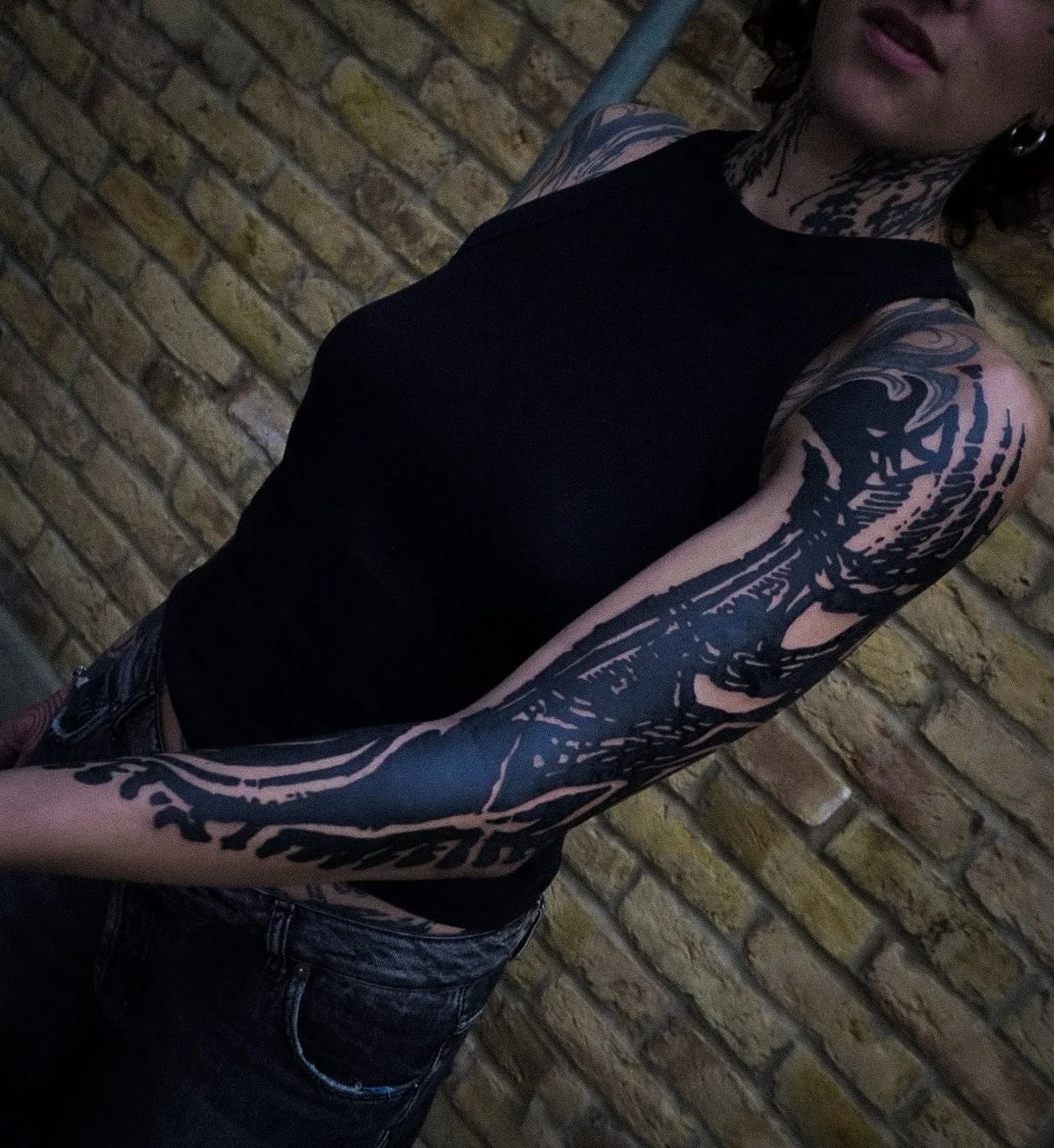 A person with tattoos on their arm and neck, wearing a black sleeveless top and dark jeans, standing against a brick wall.