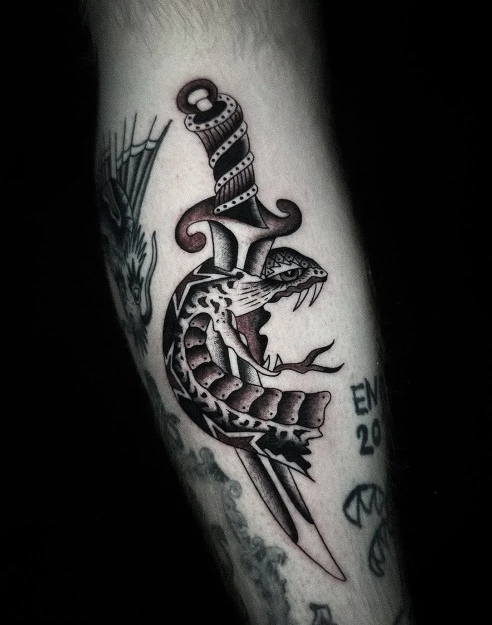 Tattoo of a snake with an open mouth, tongue out, and a sword piercing through it. The snake has a detailed head and body, with tribal patterns and an eye. There is also a dagger with a decorated handle passing through the snake's body.