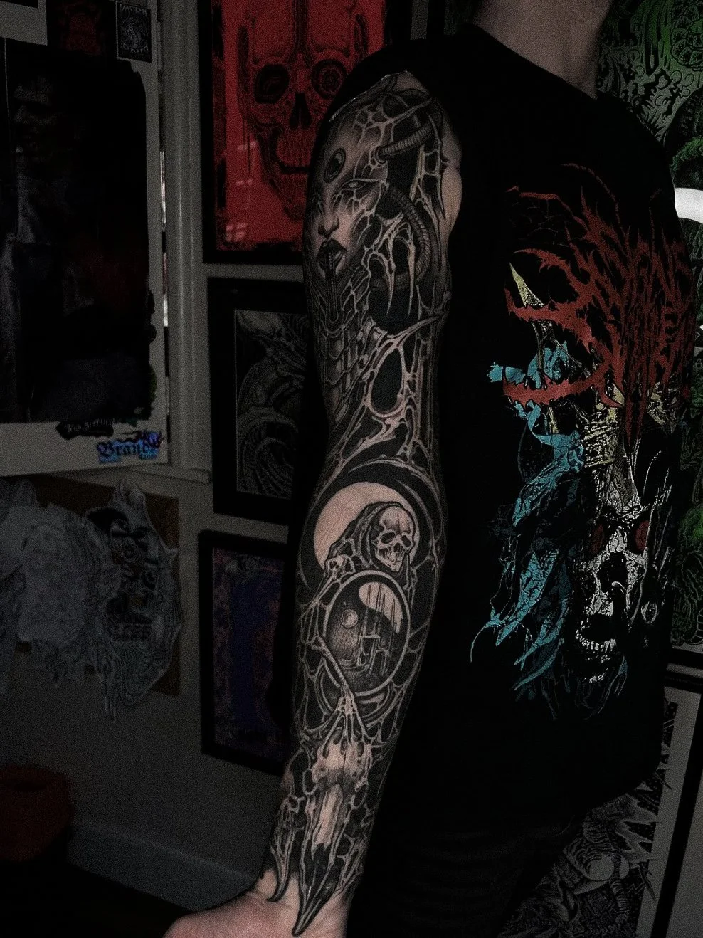 A person with a black t-shirt featuring a colorful graphic and a detailed black and gray sleeve tattoo that includes a skull and other intricate designs, standing in front of framed artwork and posters.