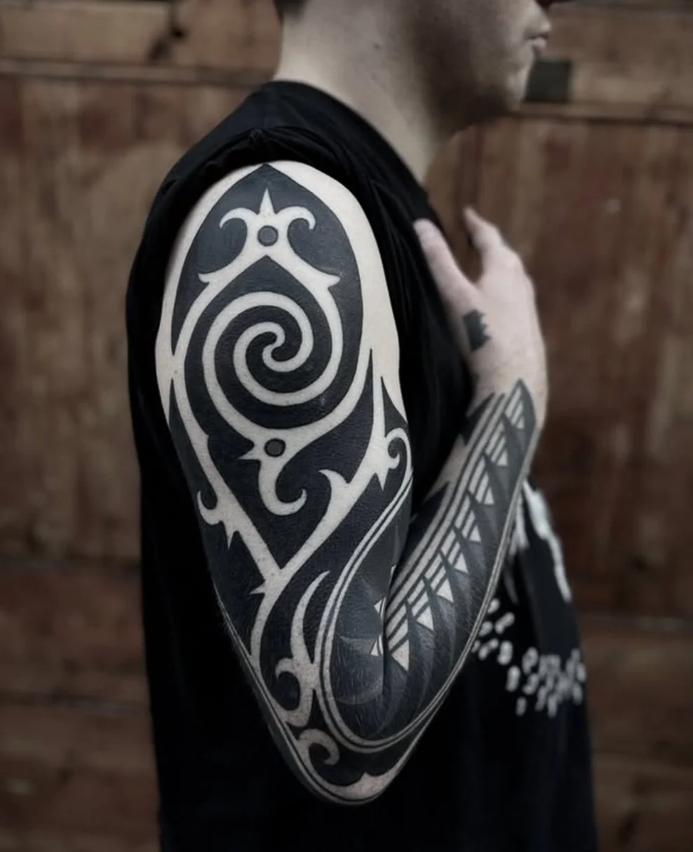 Close-up of a man's upper body showing a tribal tattoo on his left shoulder and upper arm, with his hand on his chest, against a wooden background.