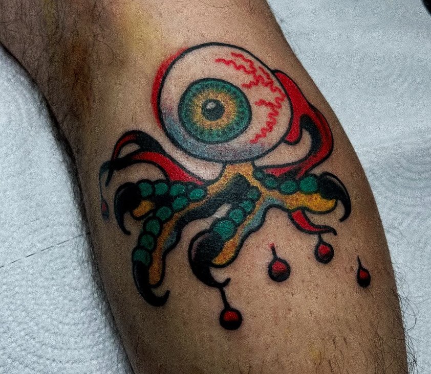 Tattoo of a cartoonish eyeball with a rainbow-colored iris, attached to a stylized octopus with green and yellow tentacles, on a person's arm.