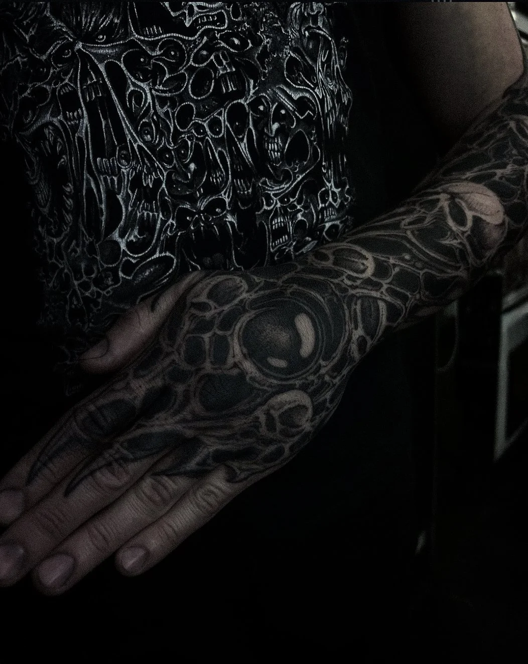Person with extensive black and gray tattoos covering their arm, hand, and chest, wearing a black shirt with white skull and abstract designs.