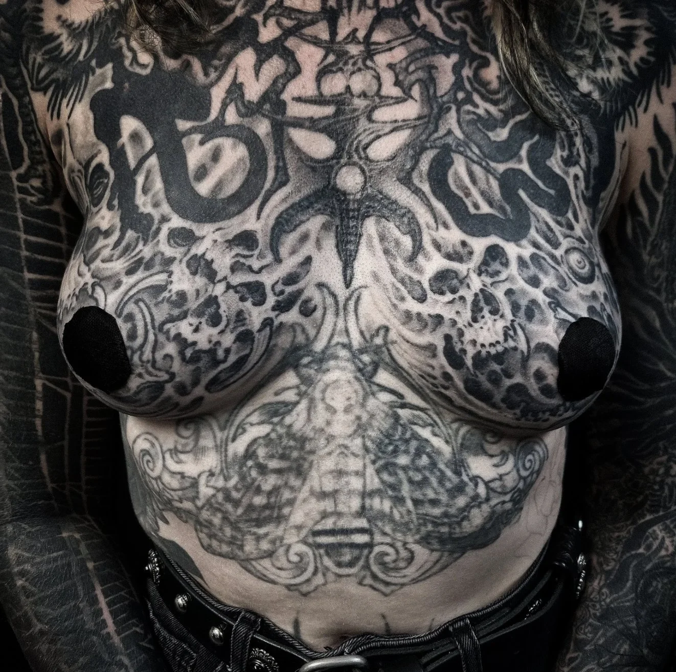 Close-up of a person's chest and abdomen covered in black tattoos, including a large dragon, skulls, and various intricate designs.