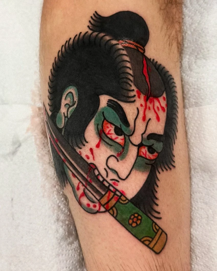 Tattoo of a fierce-looking samurai with black hair, green makeup around the eyes, holding a katana sword with a decorated handle, and blood splatters on the face and neck.