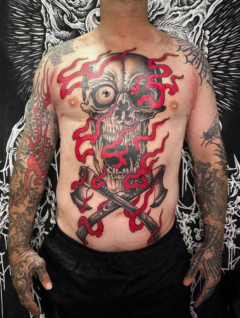 A person with tattoos on both arms and a large tattoo on their chest, featuring a flaming skull with crossed axes below it, in a dark and intricate background.