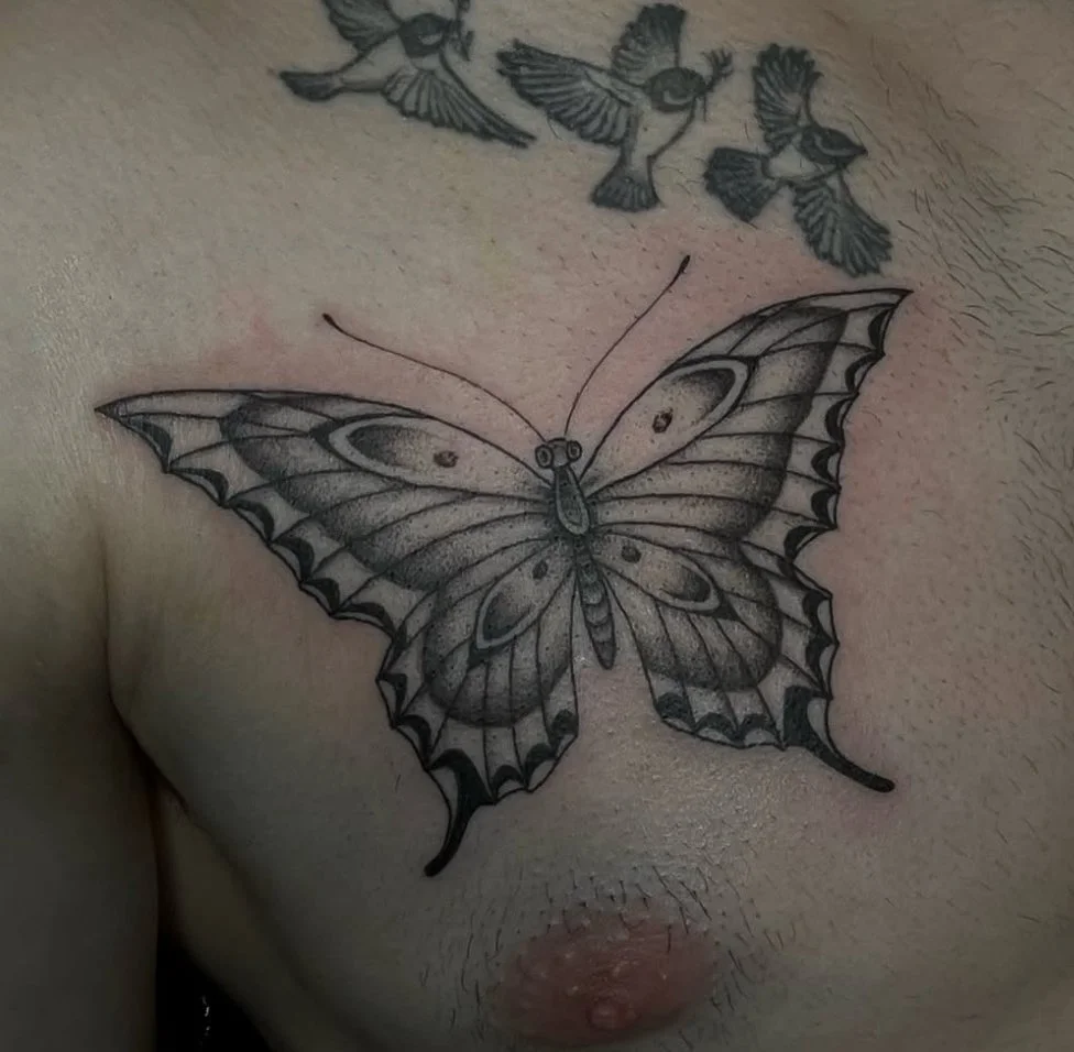 Black and gray butterfly tattoo on person's chest with smaller bird tattoos above.