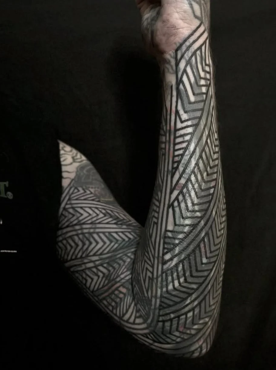 Close-up of a person's arm and shoulder with intricate black and white geometric tribal-style tattoos against a black background.