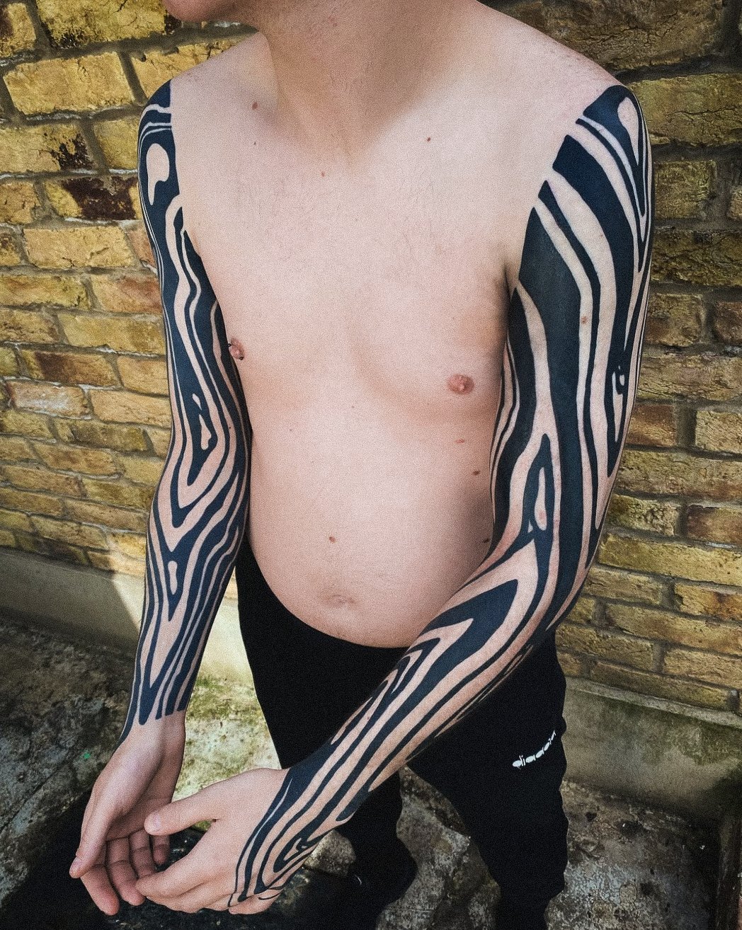 Person with tattoos on both arms, shirtless, standing outdoors against a brick wall.