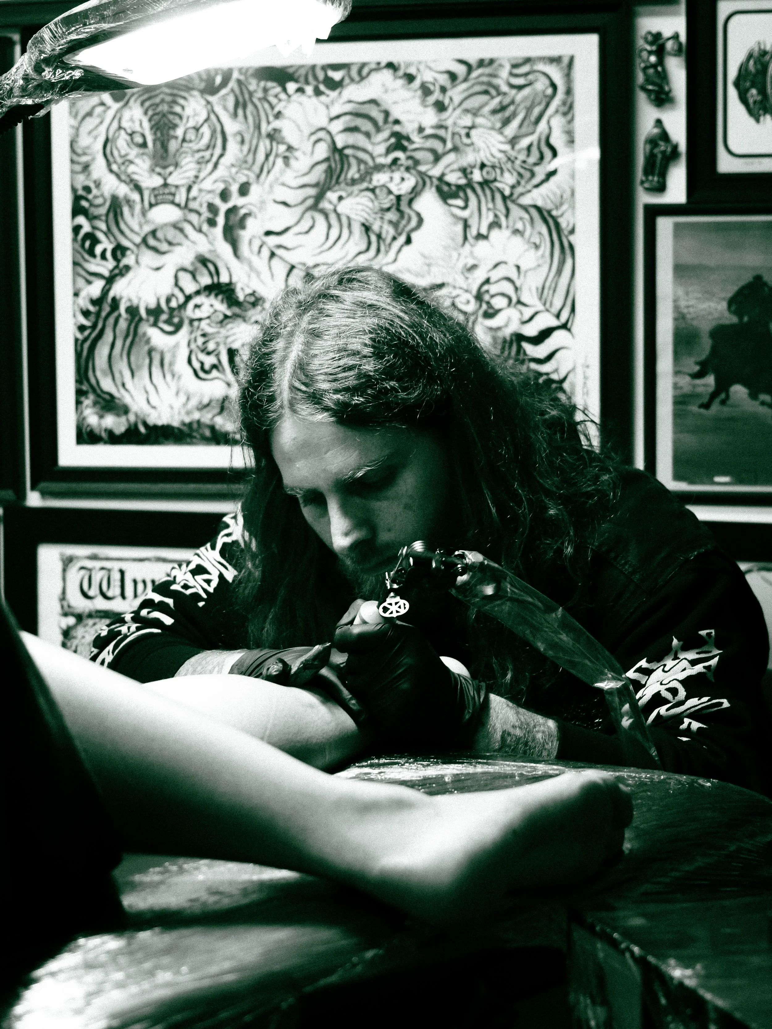 Tattoo artist with long hair and black gloves working on a client's arm in a tattoo studio decorated with framed artwork.
