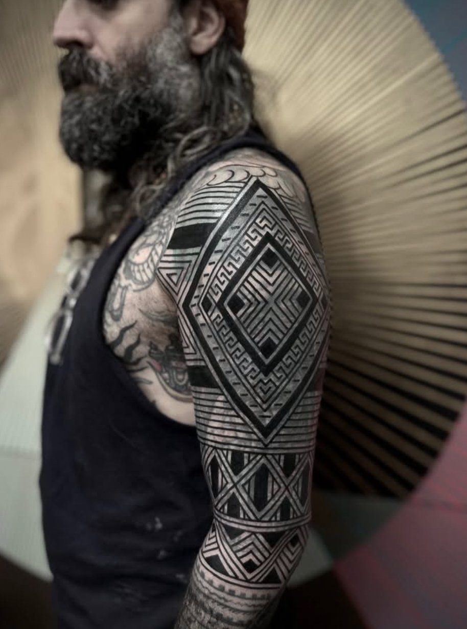 A man with long hair and a beard showing an intricate black and gray geometric tattoo on his upper arm and shoulder.