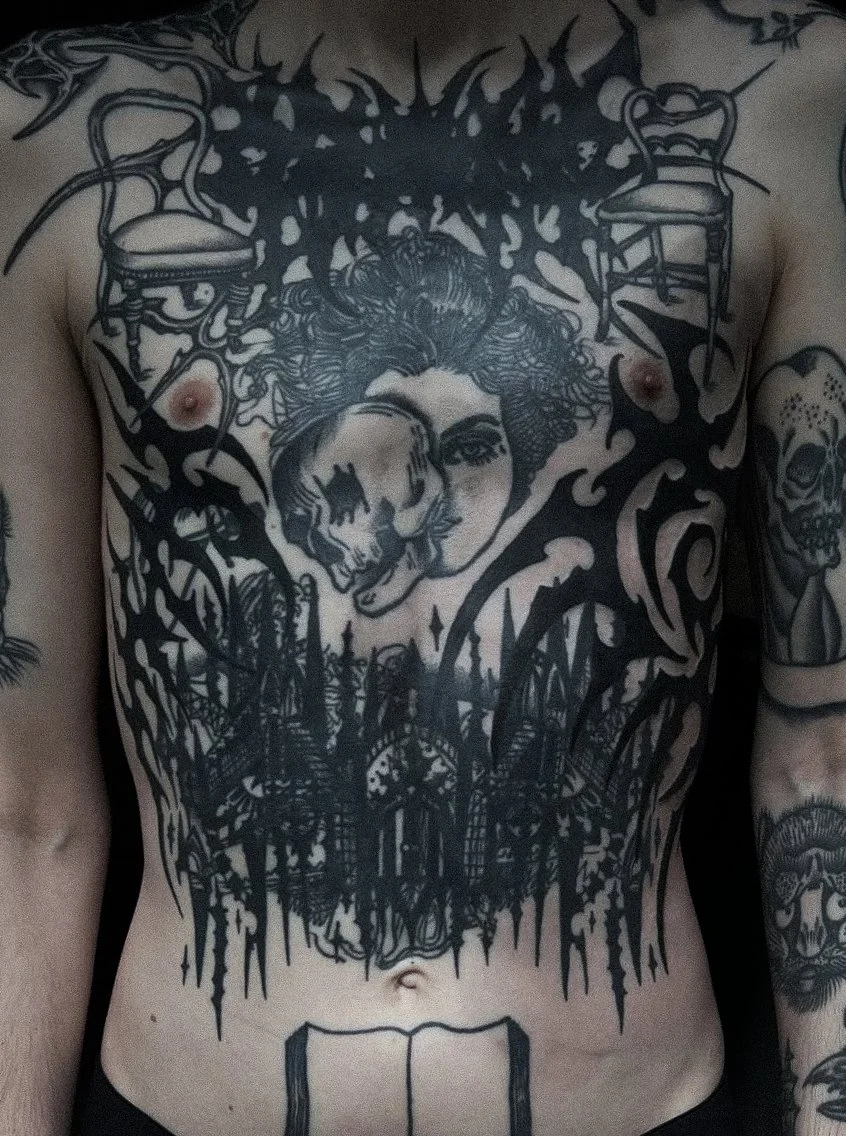 A person's chest with multiple black tattoos, including a large face of a woman, a skull, chairs, flames, and various abstract designs.