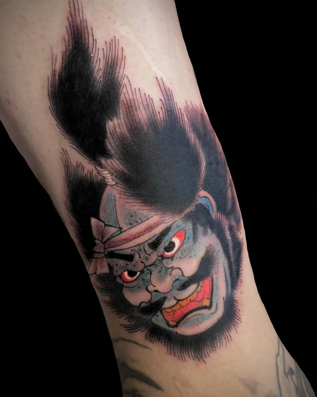 Tattoo of a fierce-looking Japanese demon or ogre with an angry expression, sharp teeth, and red eyes, featuring traditional Japanese tattoo style with bold lines and dark shading.