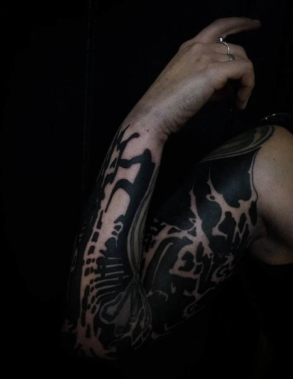 A close-up of a person's arm with a black tattoo, holding their hand near their face against a dark background.