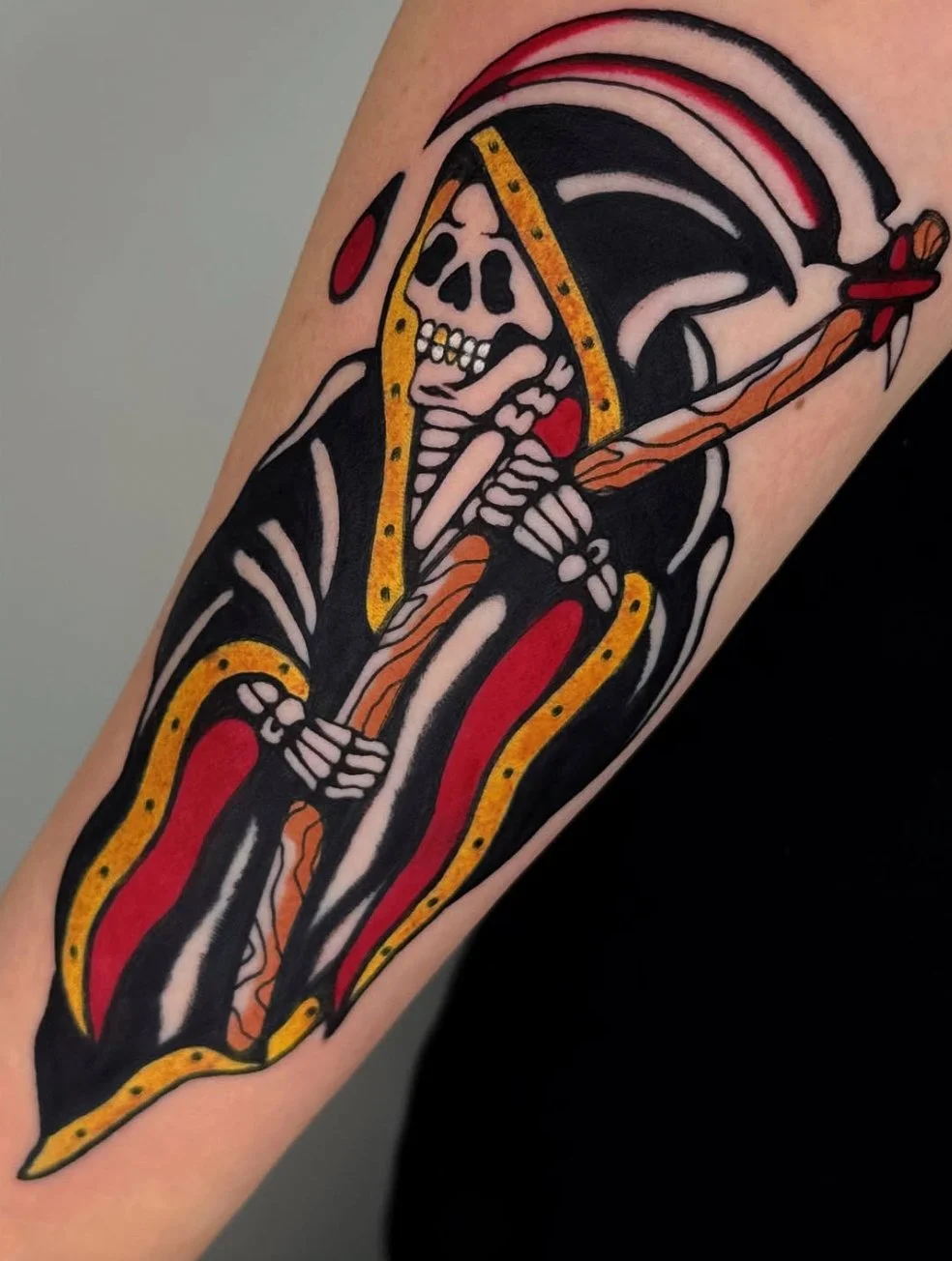 Tattoo of a skeleton wearing a hooded cloak, holding a sword, with bold black, red, yellow, and orange colors.