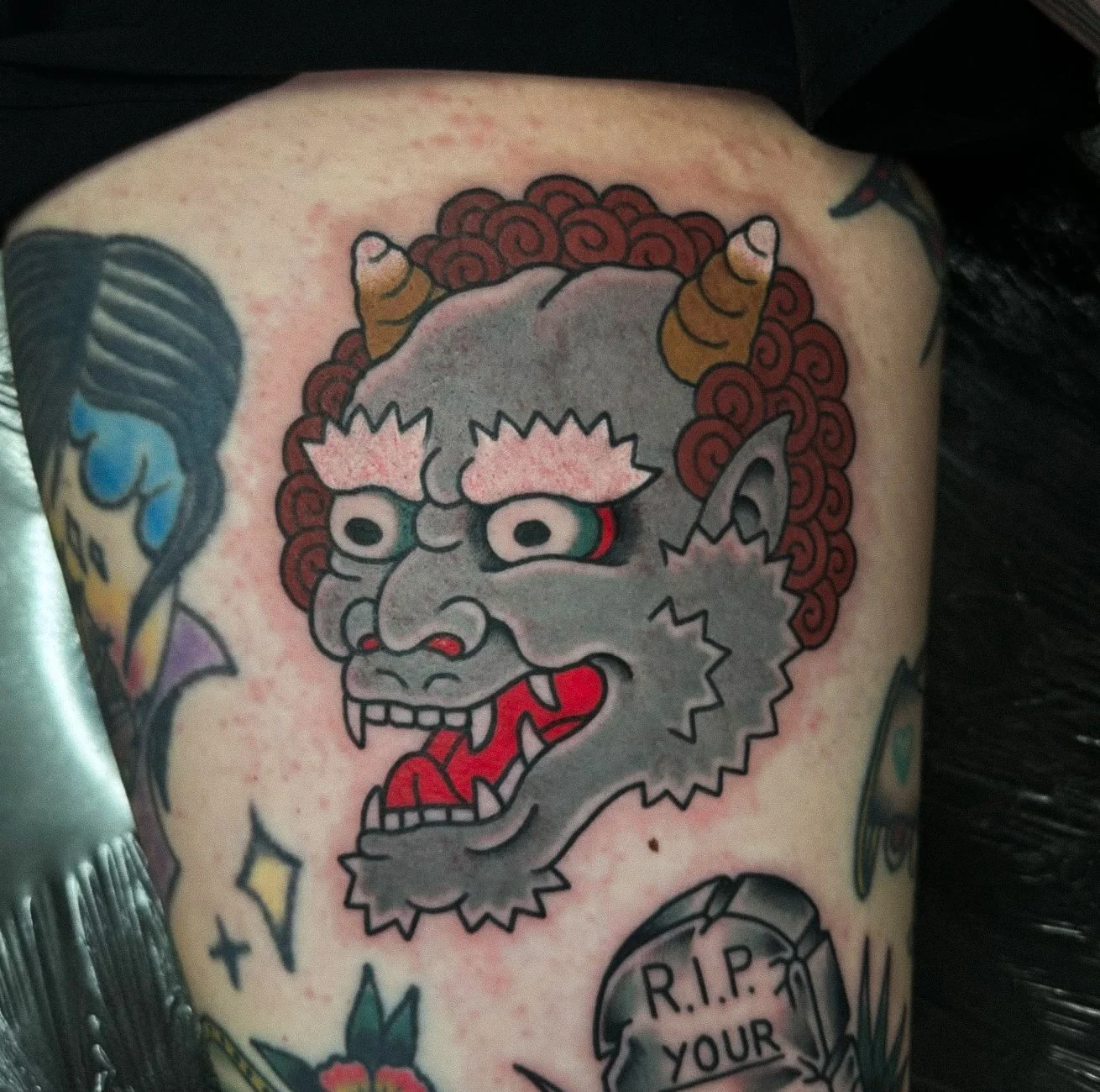 Traditional Japanese style oni mask tattoo on person's shoulder, depicting a fierce demon with gray face, sharp teeth, red tongue, and prominent horns surrounded by red curly hair.