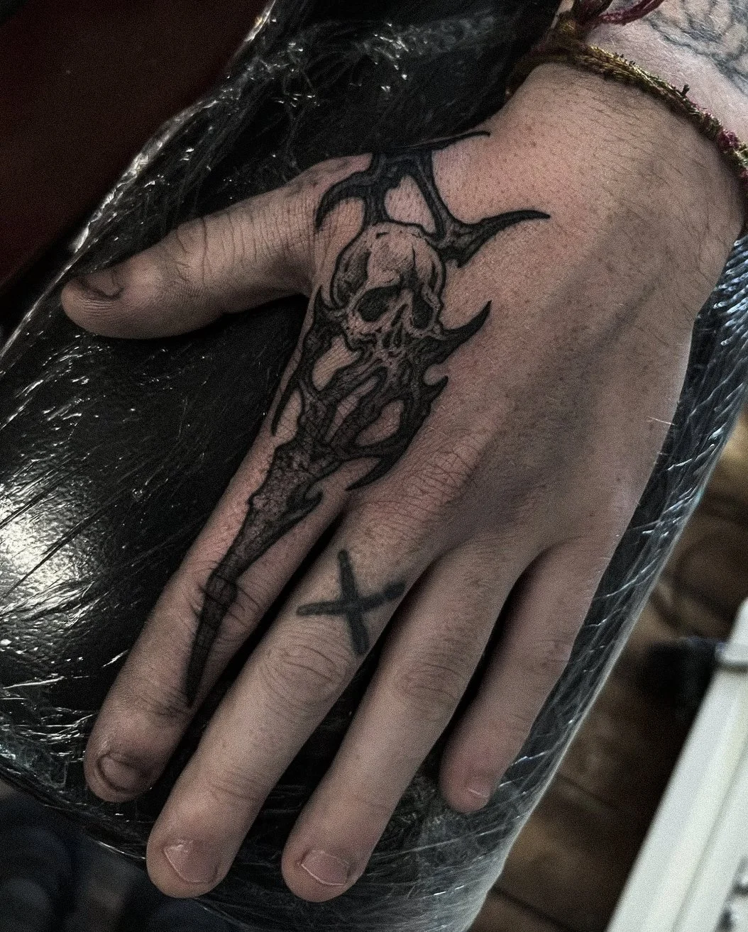 Black ink tattoo of a skull with horns and wings on the top of the hand, extending toward the wrist, with a small 