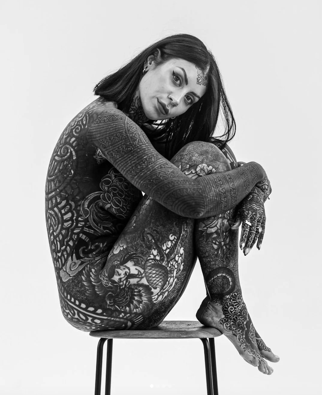 Black and white photo of a woman with extensive tattoos sitting on a wooden chair, hugging her knees, looking at the camera.